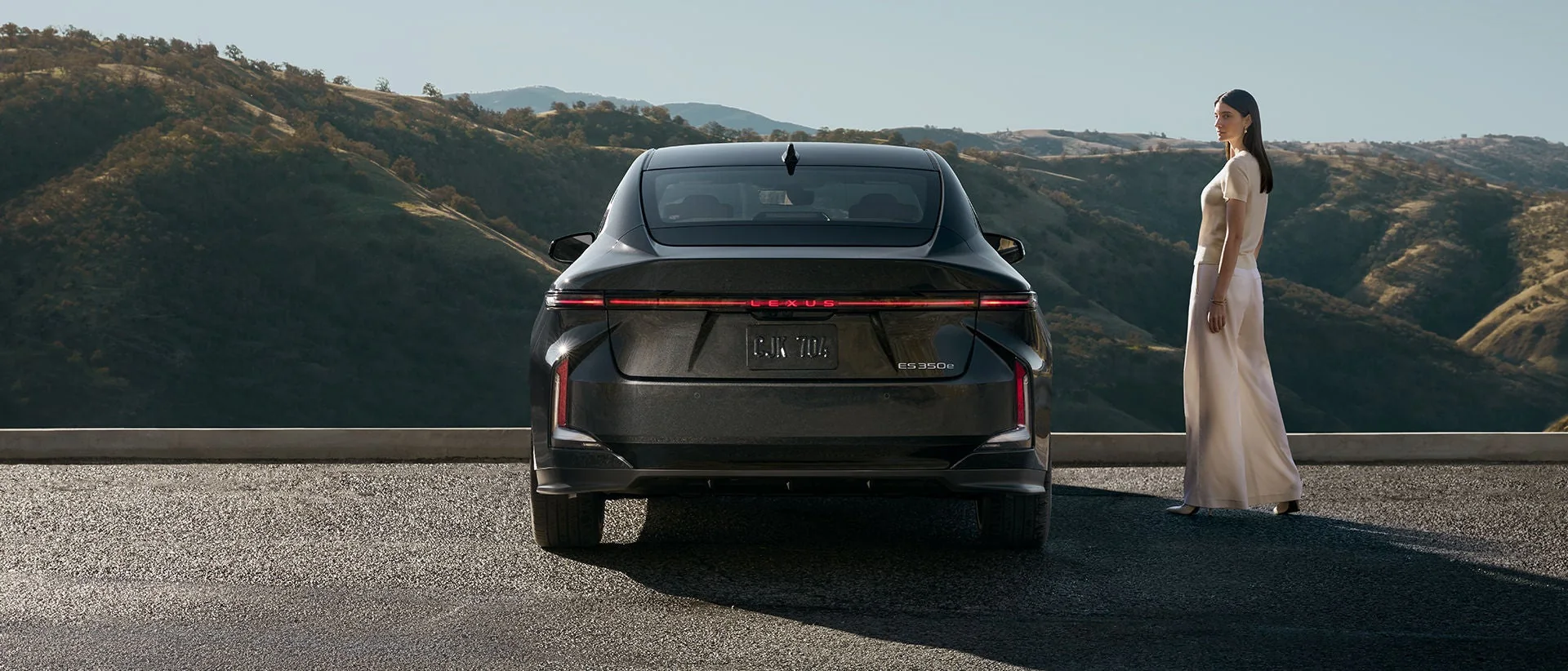A black Lexus ES seen from behind at a hillside overlook, a woman in white standing beside it