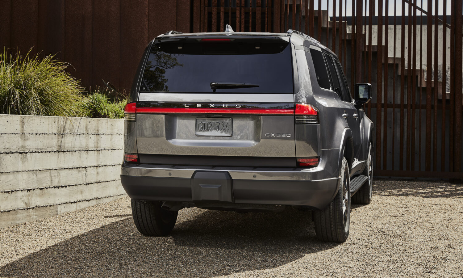 A dark gray Lexus GX Premium parked on gravel beside a timber fence, seen from behind