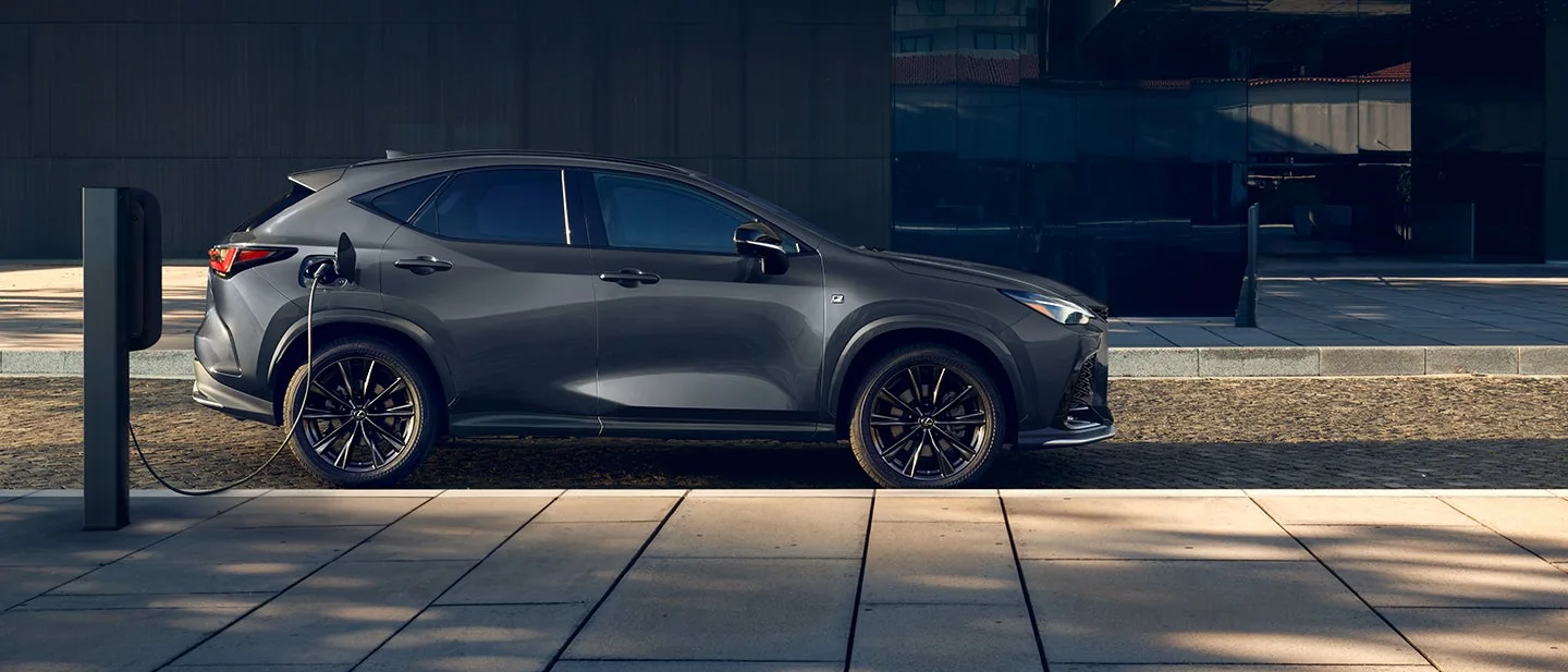 A dark gray Lexus NX plug-in hybrid charging at a post beside a glass building