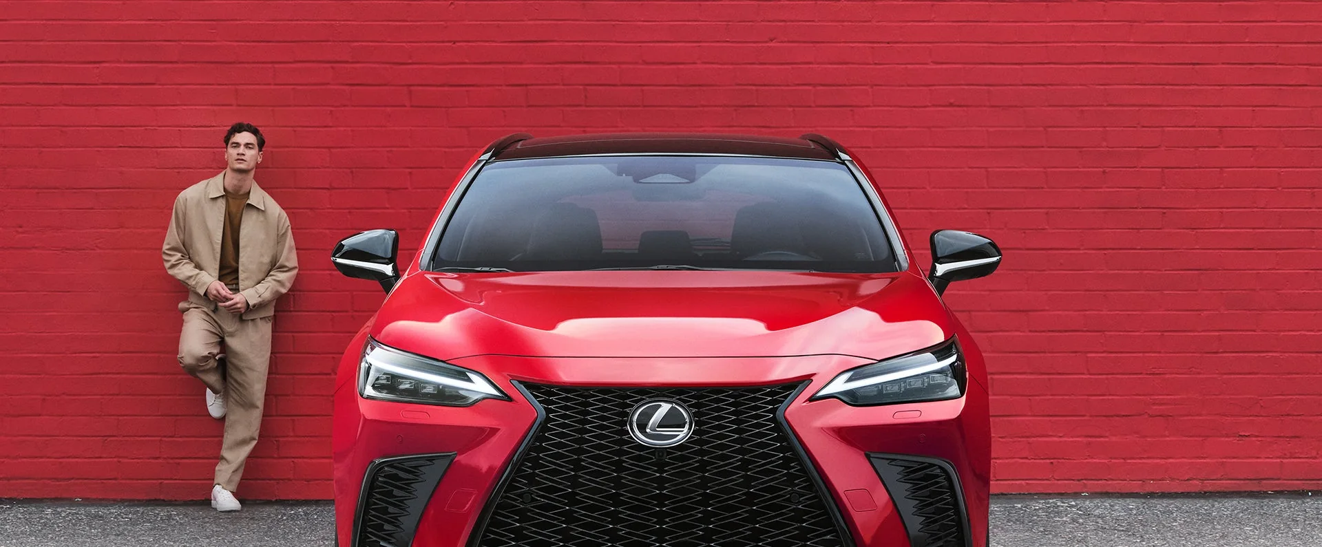 The front of a red Lexus NX parked beside a red brick wall, a man leaning on the wall next to it